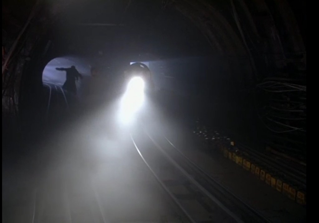 A subway tunnel in which an oncoming subway glares with a white light in the darkness. A figure flees. 